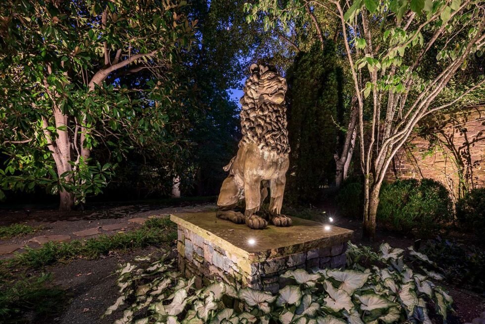 Lion statue situated within a garden is lit with accent lights. Landscaping lights also illuminate the surrounding trees and brick structure in the background.