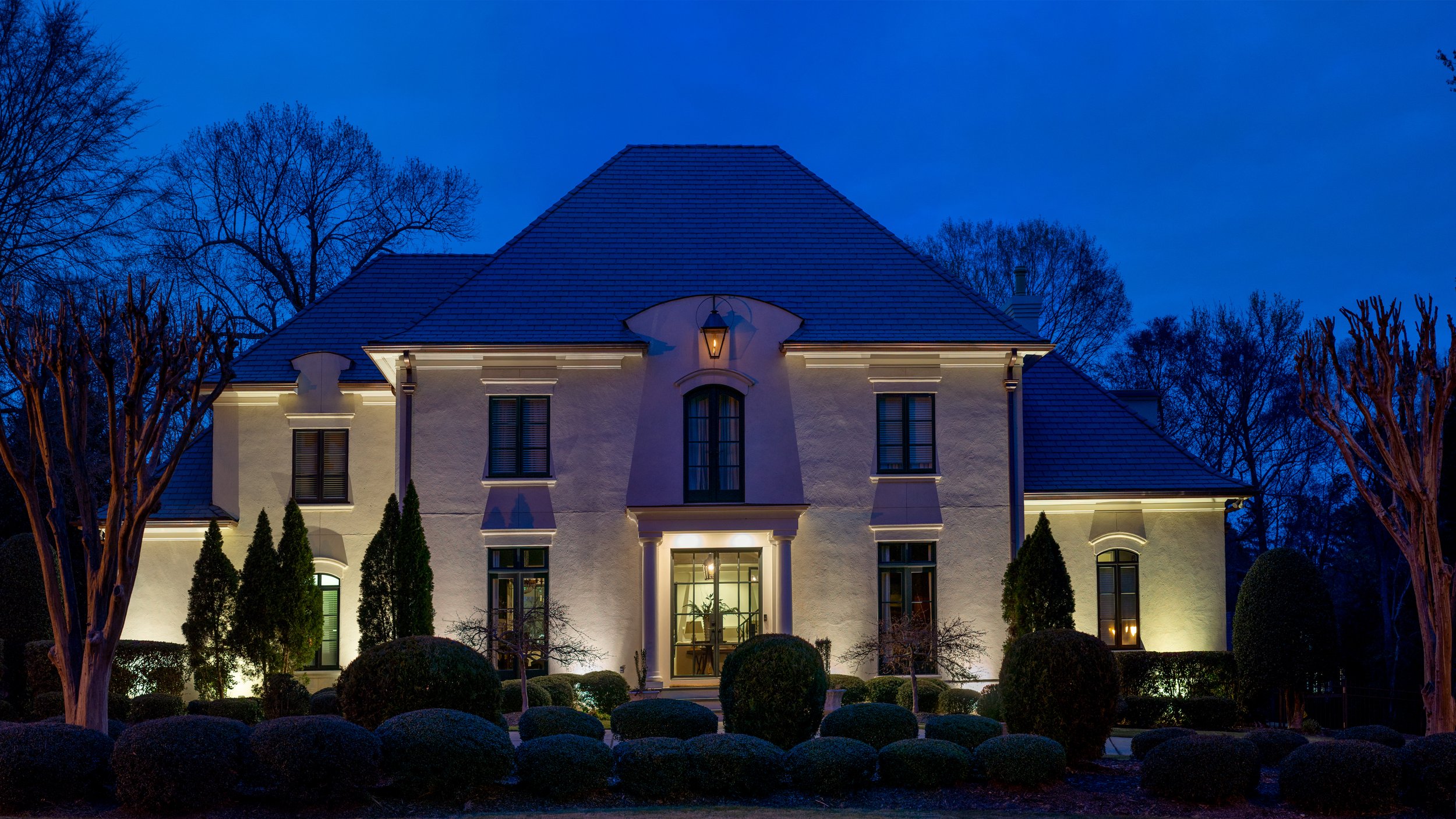 Architectural landscape lighting illuminates a large white home with silhouetted trees.
