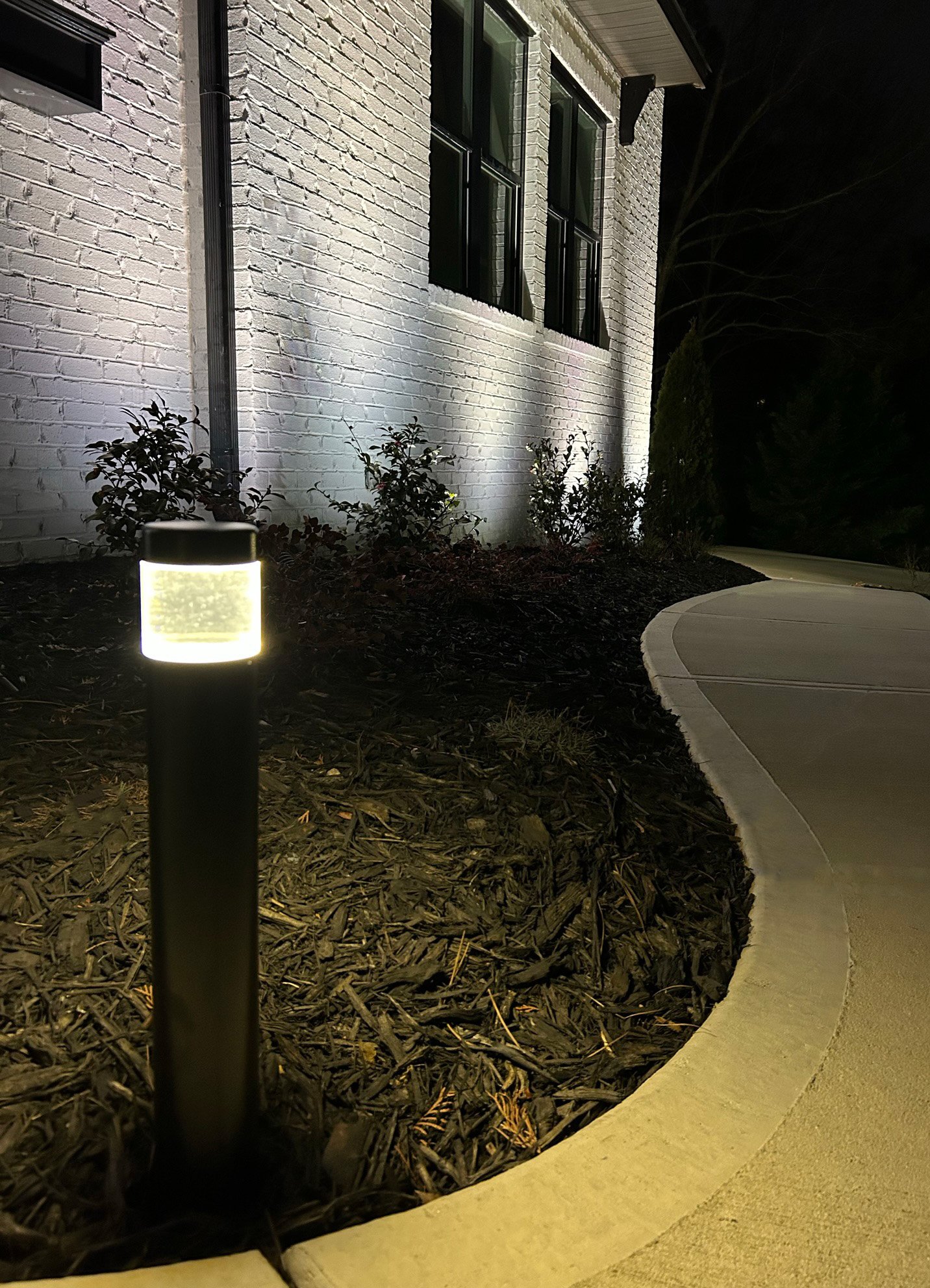 Bollard style path light illuminates the front path leading up to a white brick home with architectural up lights.