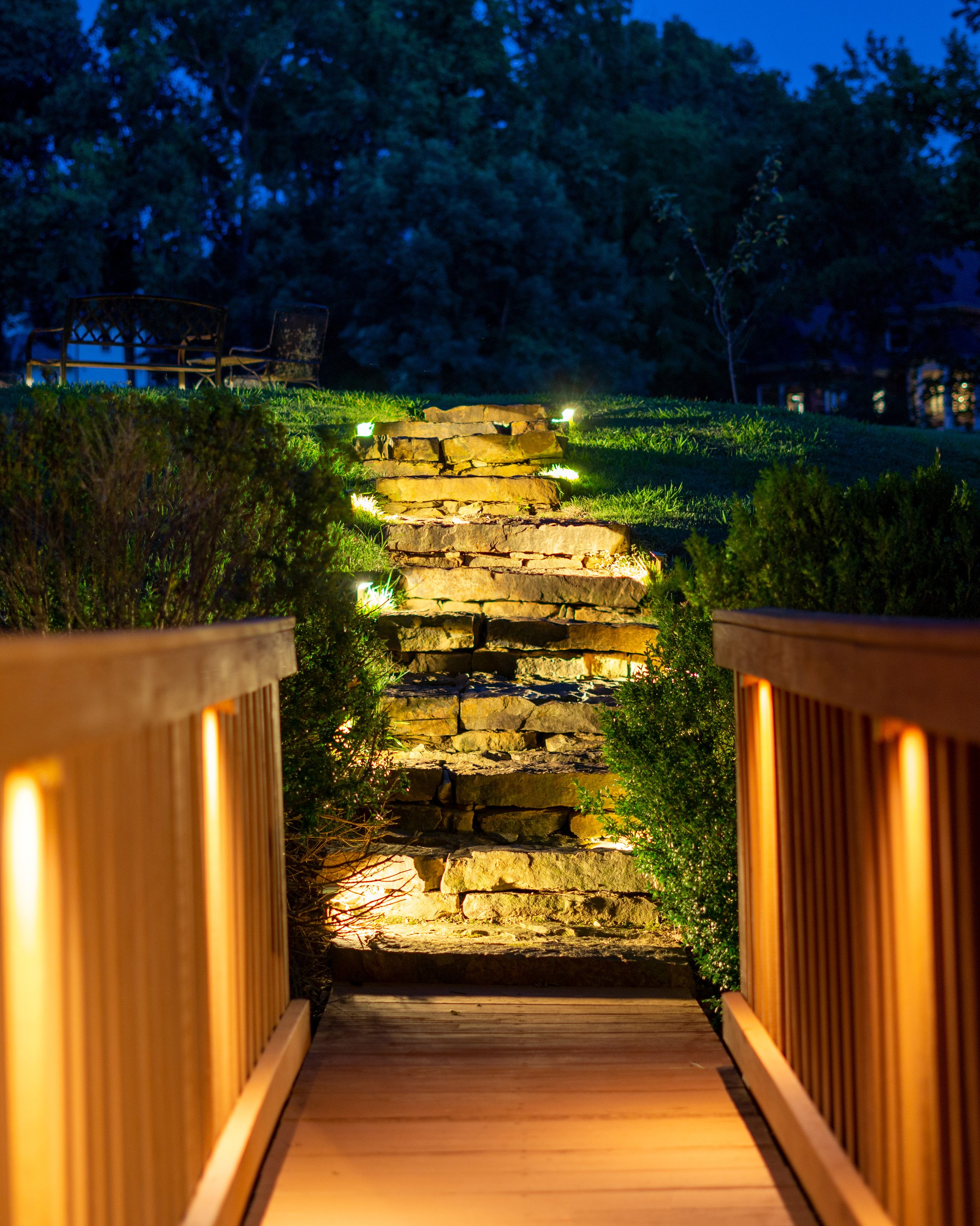 An orange colored wood bridge illuminated by undermount deck lights leads the way to a natural stone path illuminated with low profile path lights.