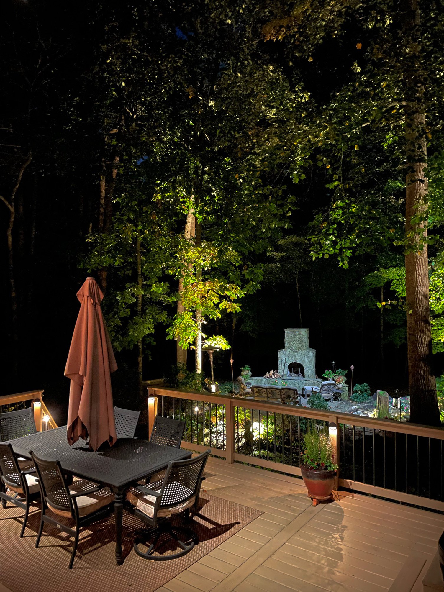 Overhead deck lighting illuminates an outdoor eating area. In the background you can see additional landscape lighting on the trees and outdoor fireplace.