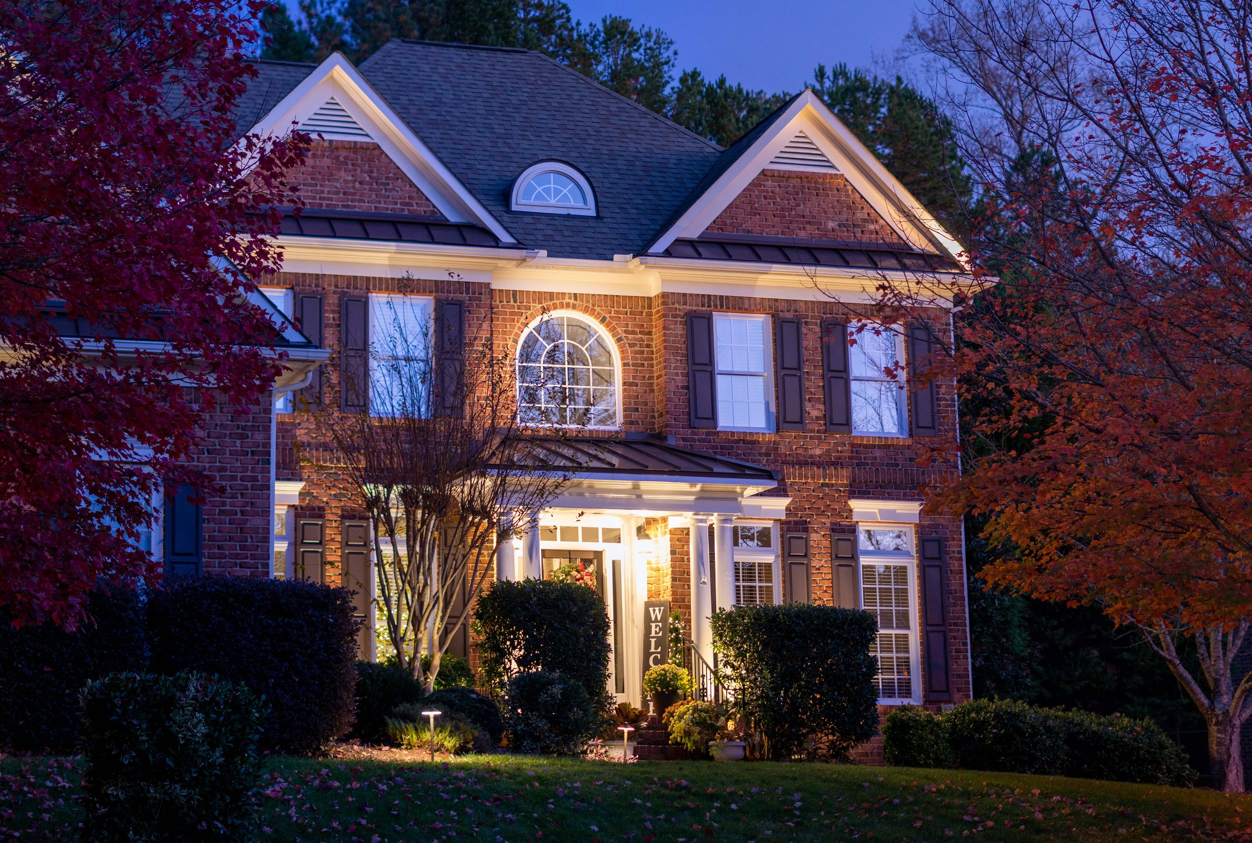 Traditional brick home with shutters illuminated with architectural up lights and gutter mount landscape lighting.