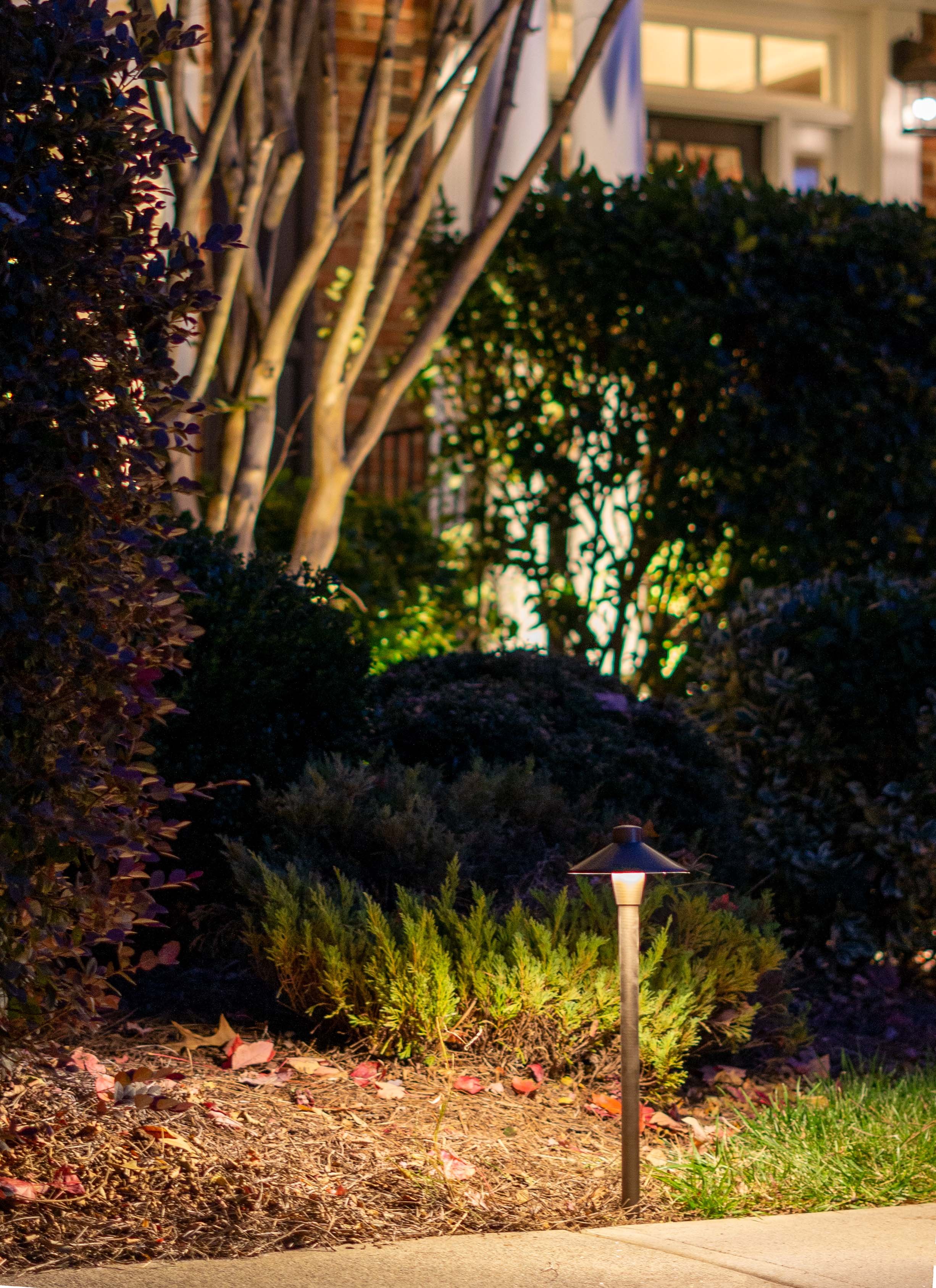 Close up image of a traditional path light at the edge of a landscaping bed. Plants and trees can be seen illuminated with landscape lighting in the background.