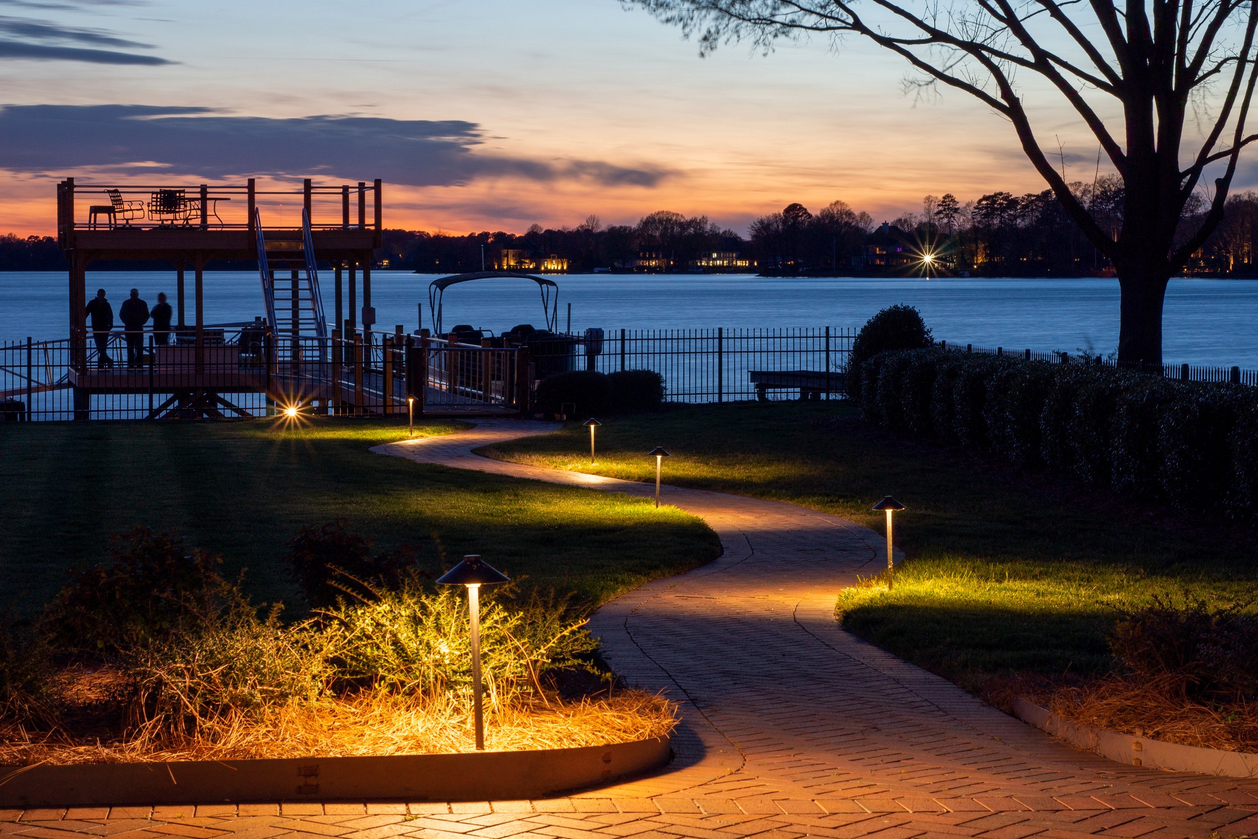 Winding path with staggered path lights leads down to a silhouetted tree, dock, and boat. A beautiful lake and sunset sky is visible.