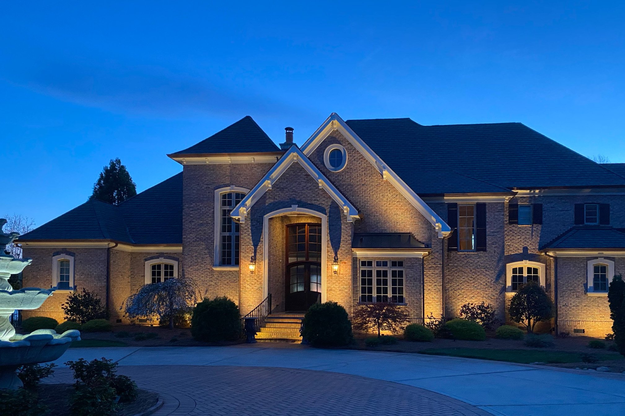 Large, light colored brick home with architectural up lighting and landscape lighting.