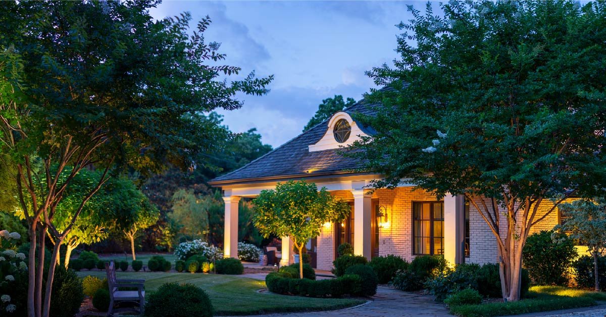 Landscape lighting and architectural up lighting illuminates a white brick home with four large columns.
