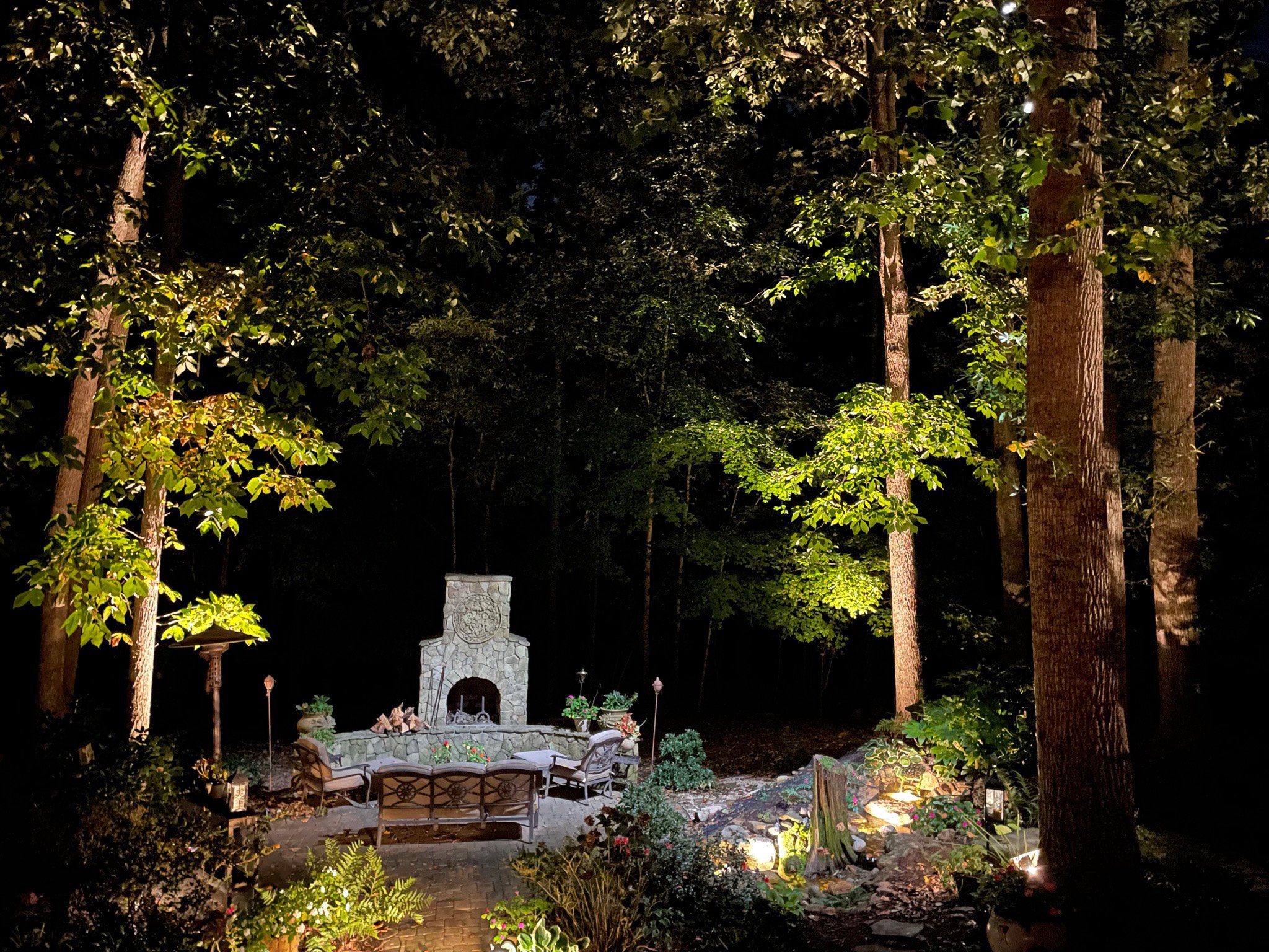 Wooded lot with a water feature and fireplace is illuminated with landscape lighting. Tree accent lights are placed at the base of larger trees framing the scene. A cool colored down light gently illuminates the sitting area.