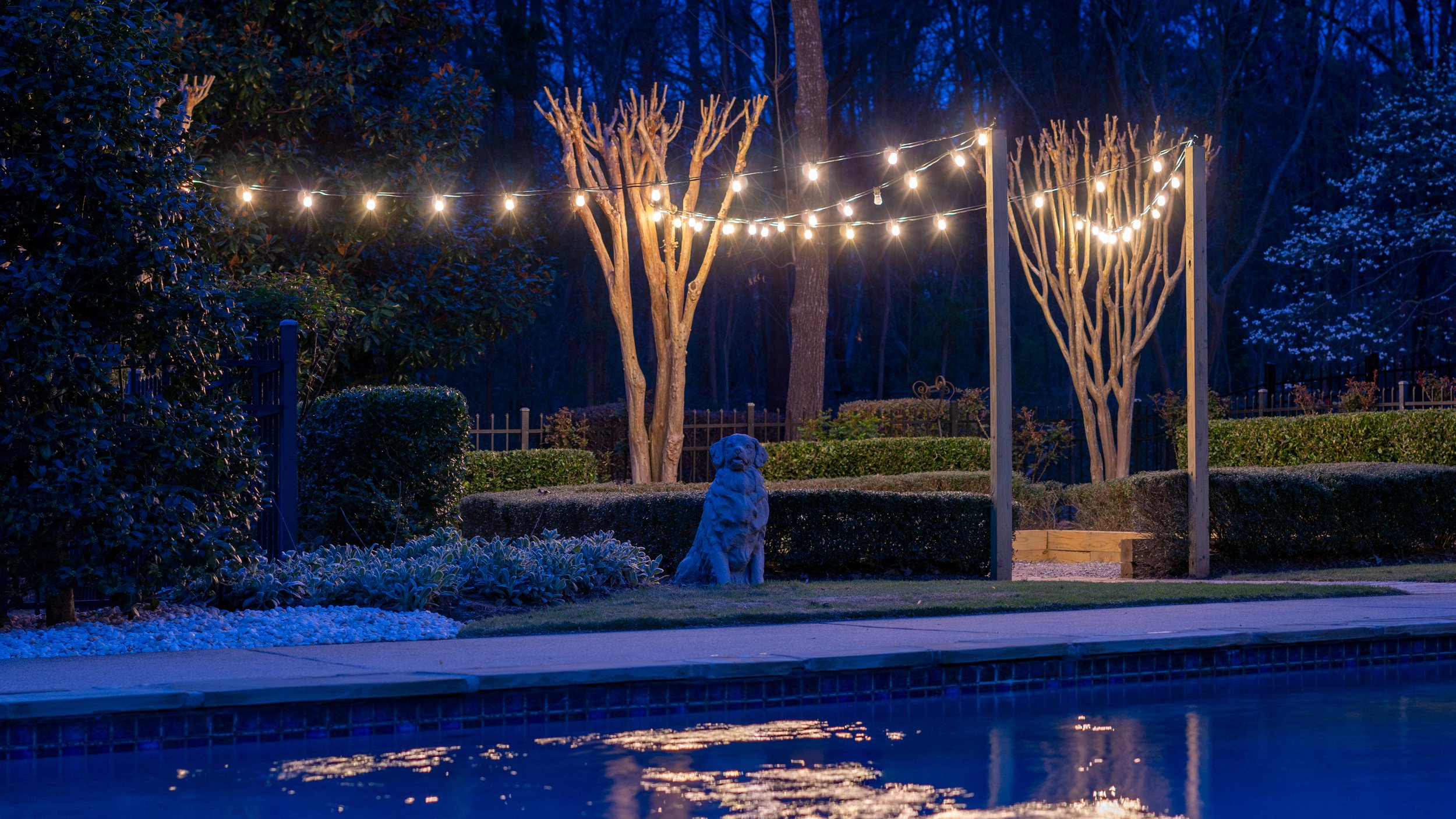Bistro lights strung between posts and crepe myrtle trees create ambient light for a poolside firepit area.