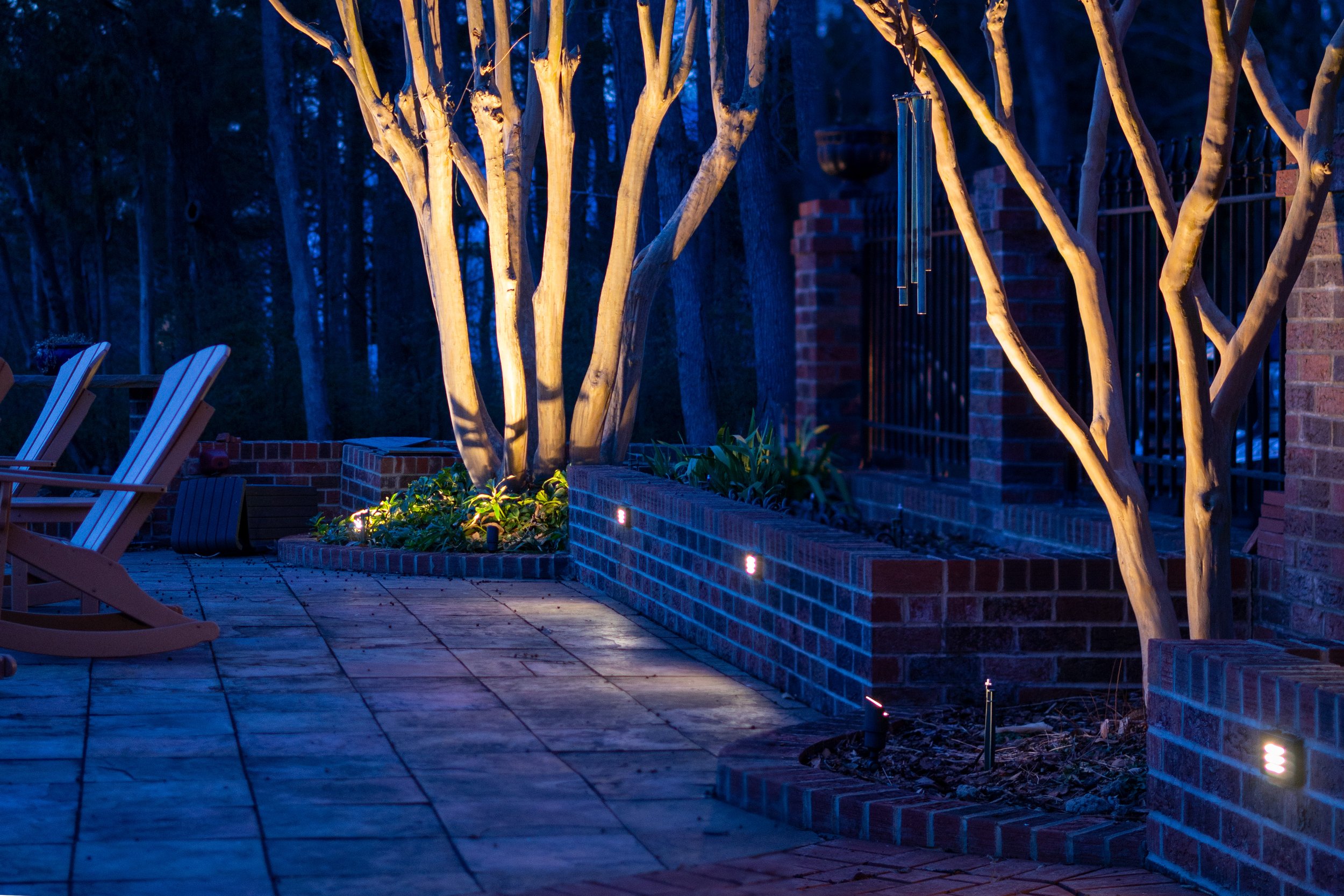 Image taken in winter of two crepe myrtle trees without leaves illuminated with accent lights. Trees are situated within a brick patio area with lights mounted in the brick to create ambient light for a fire pit area.