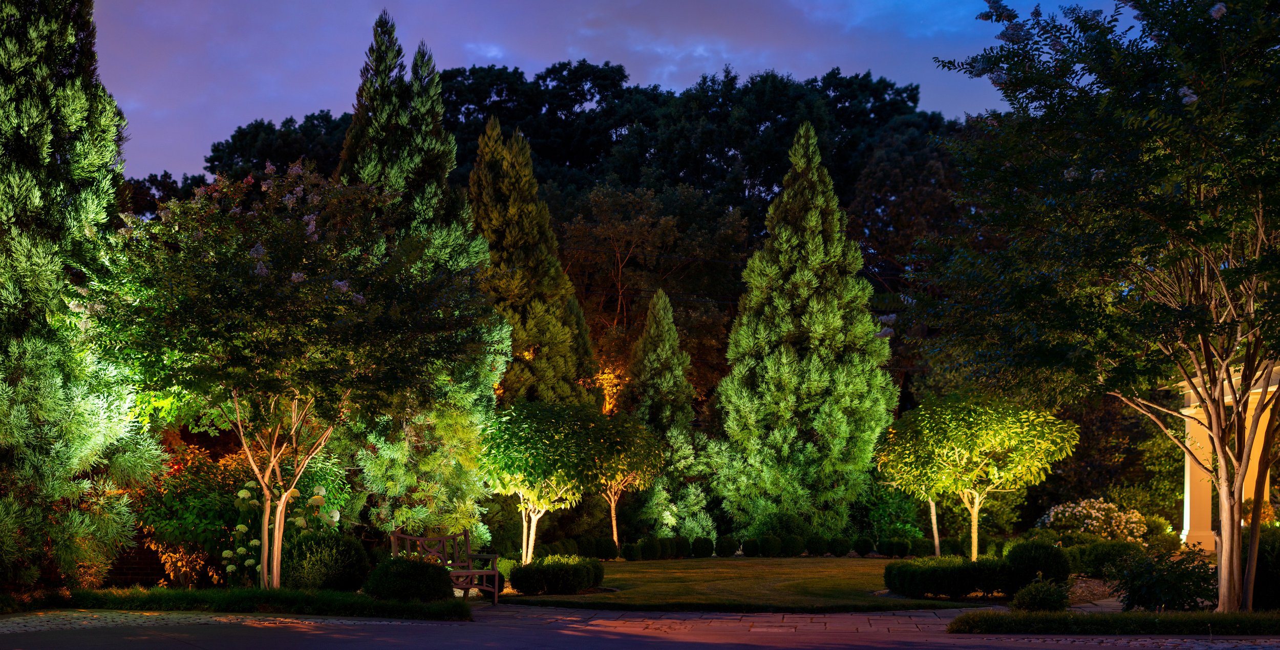Nicely landscaped front lawn with a variety of trees is illuminated with strategically placed accent lights. Landscape lighting on trees.