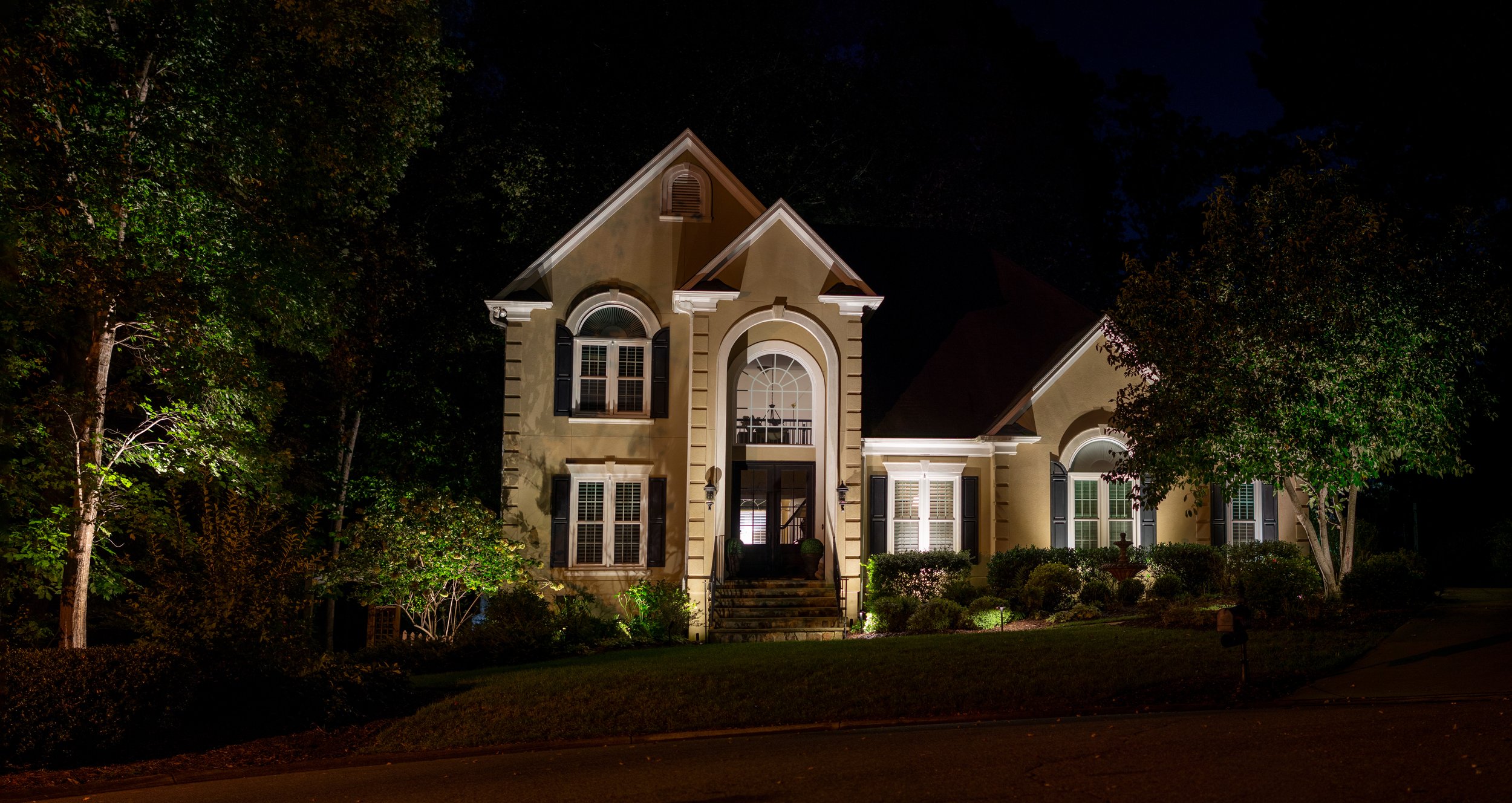 Tan stucco home with dental moulding illuminated with a combination of landscape lighting and architectural up lights.