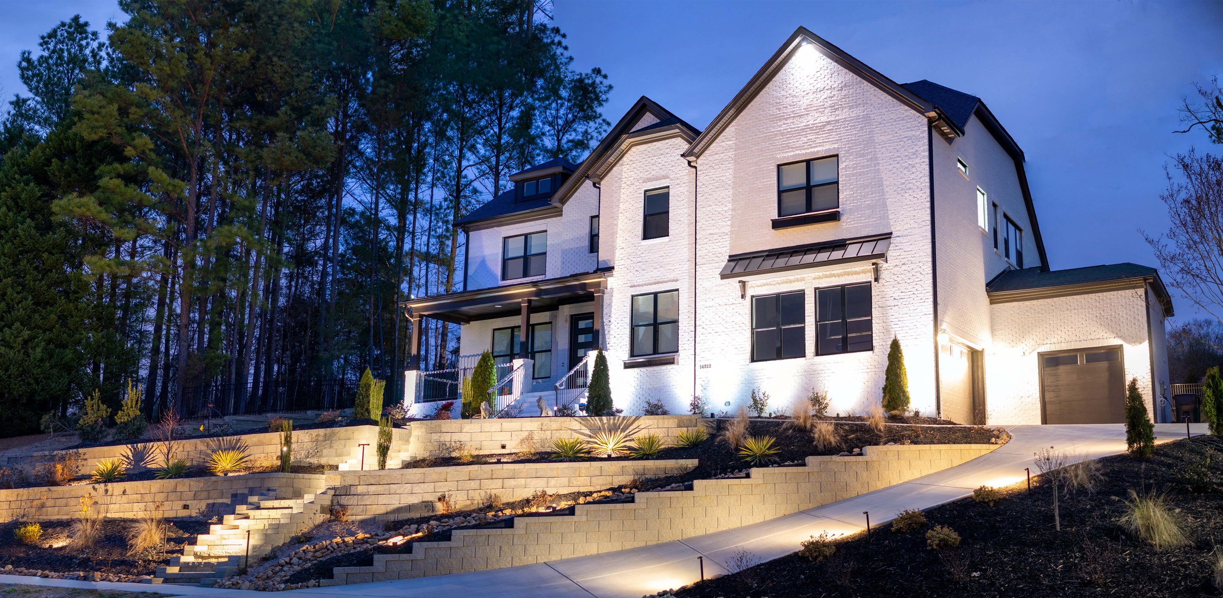 White brick home with dark trim illuminated with architectural up lights and downlights. Front yard is heavily landscaped with tiered retaining walls illuminated with soft landscape lighting.