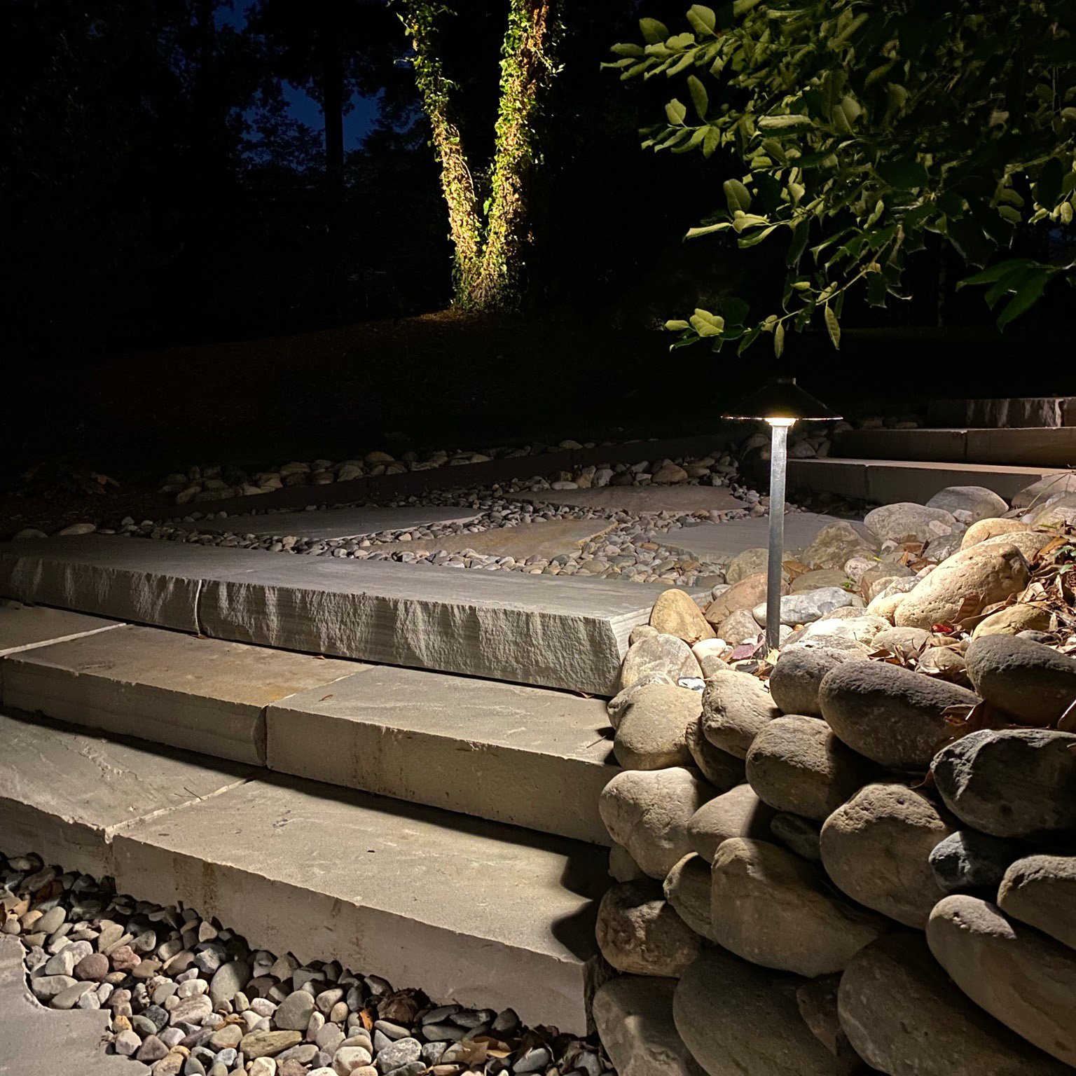 Path light illuminating stone steps surrounded by landscaping rocks. A tree with an accent light can be seen in the background of the picture.