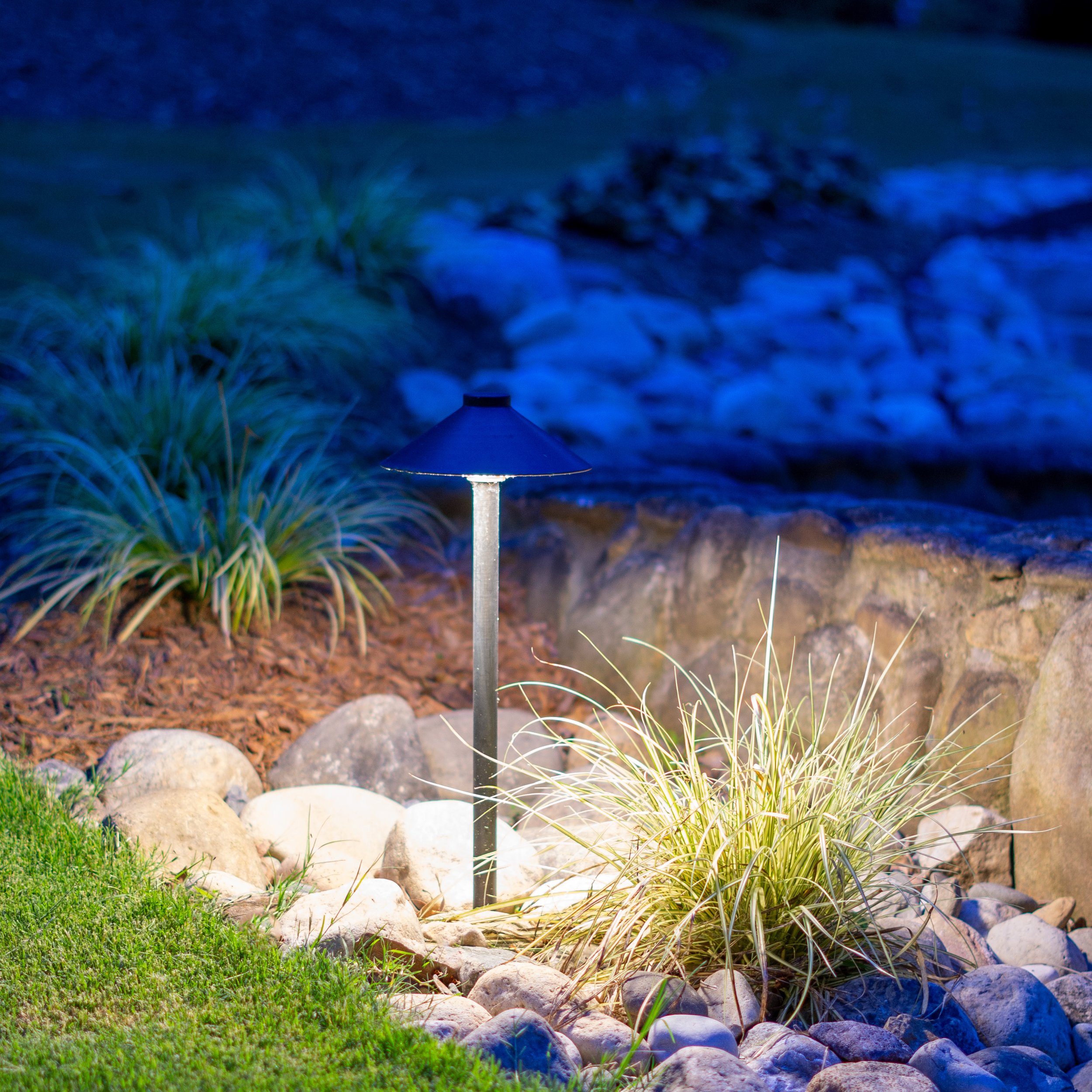 Path light used to illuminate a landscaping bed with a combination of mulch, plants, and decorative rocks.