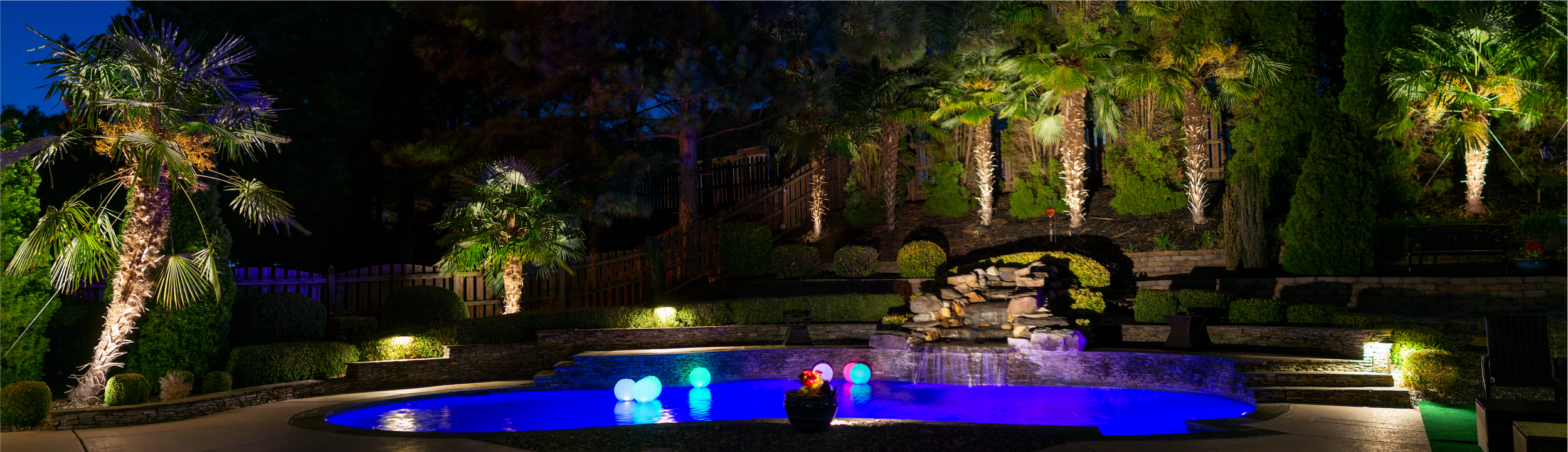 large back yard with extensive landscape lighting features a pool with an illuminated waterfall and a variety of illuminated palm trees.