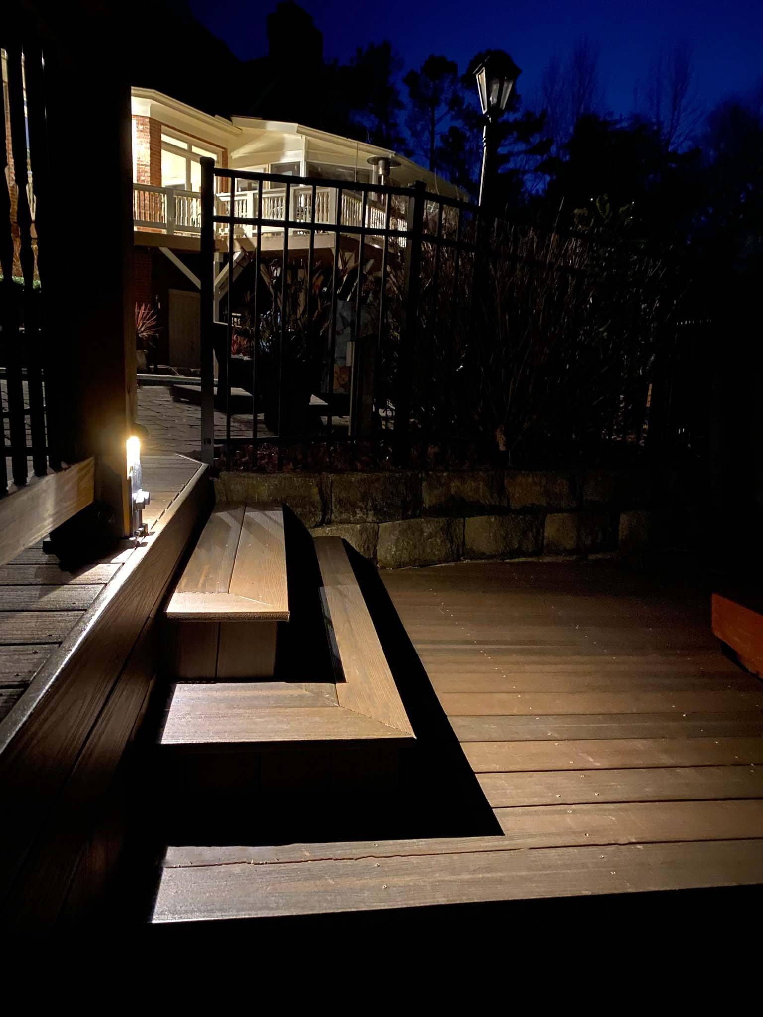 A small button deck light strategically mounted above a set of wooden steps leading from one level of a deck to the next.