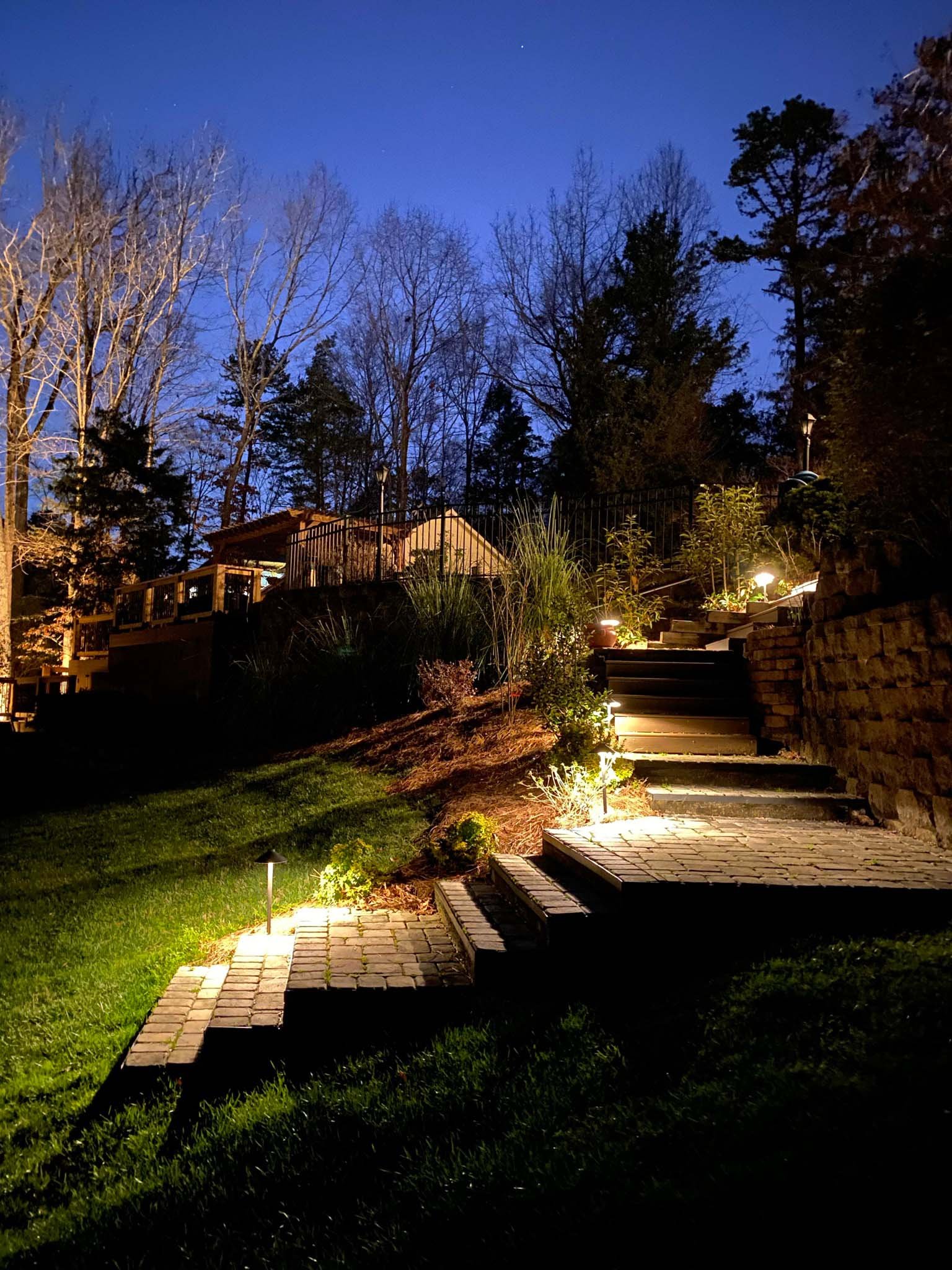 Path lights illuminate paver steps leading from a patio area down to a grassy backyard.