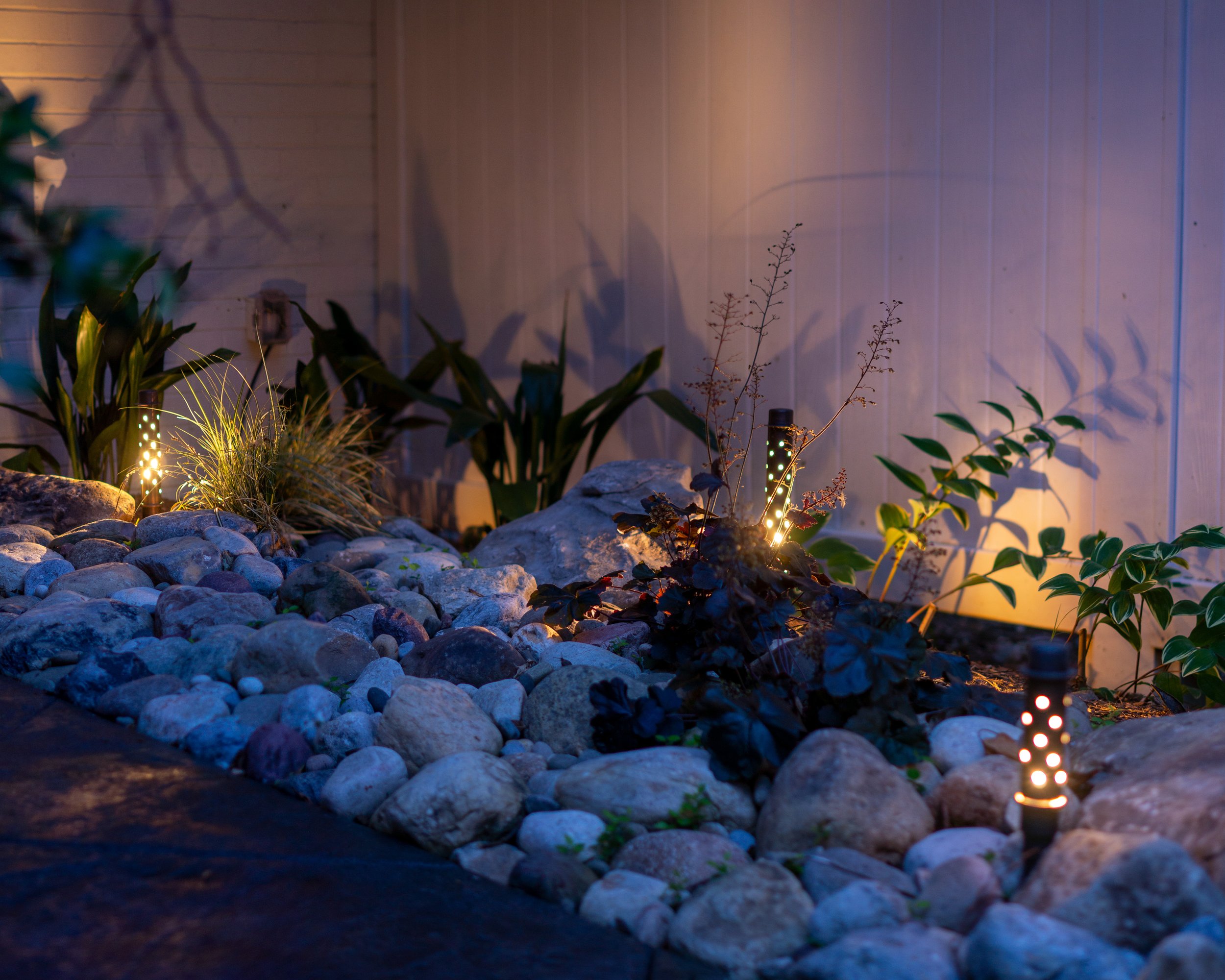 Small alcove with decorative rocks and plants is illuminated by a series of sparkler path lights creating interesting shadows on the wall.