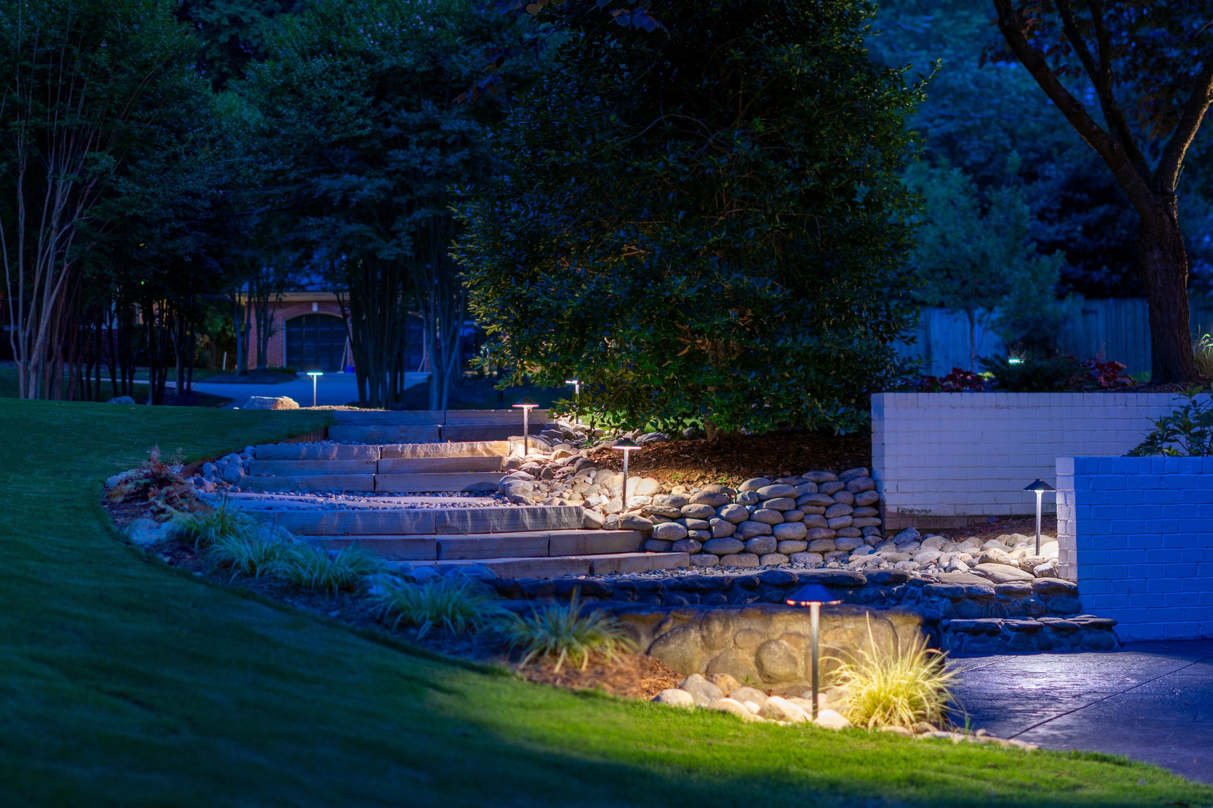Path lights illuminate natural stone steps leading from a fire pit area around the house to the driveway.