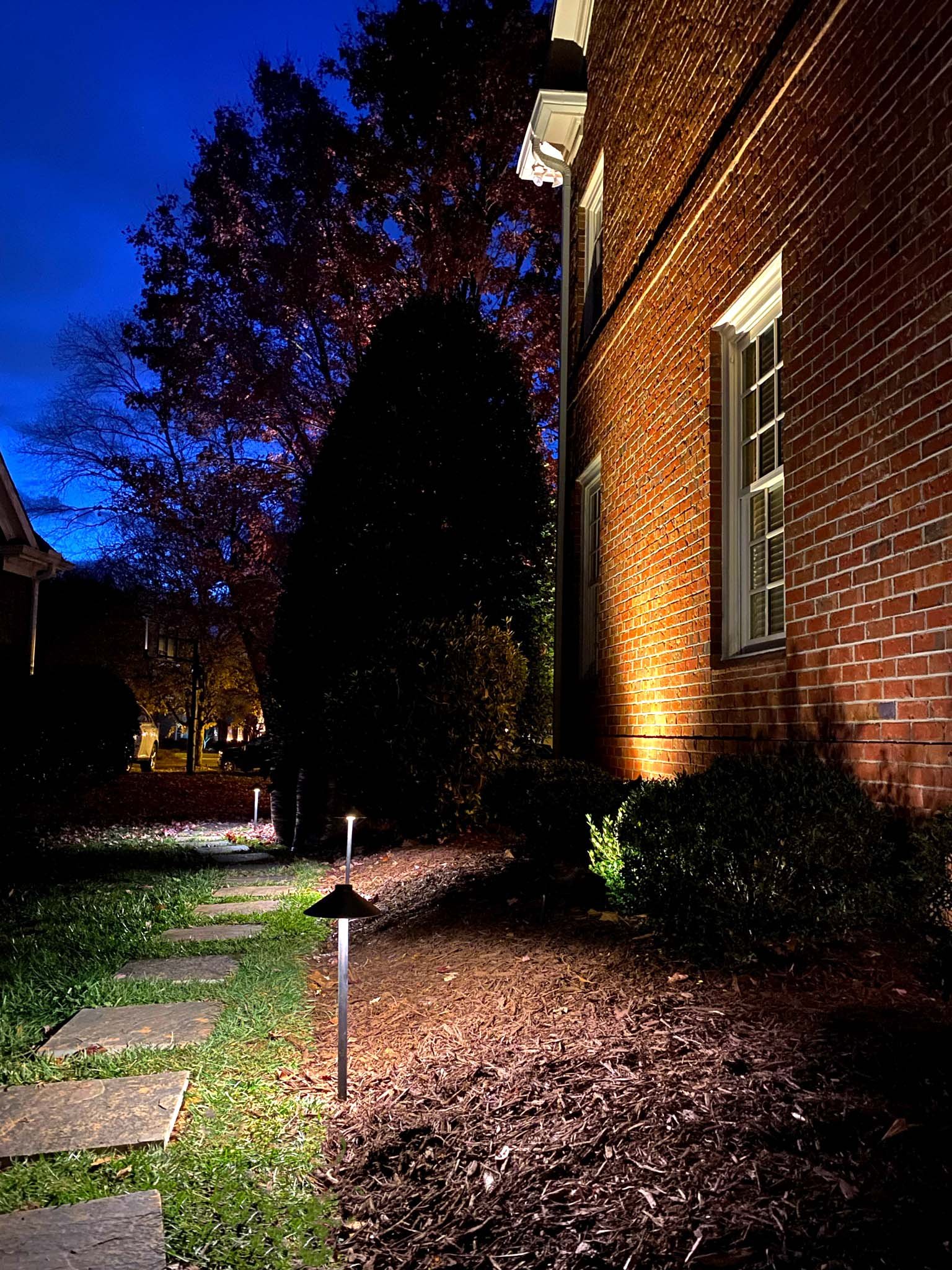 Traditional path lights illuminated a stepping stone style path on the side of a red brick home illuminated with architectural up lights and landscape lighting.