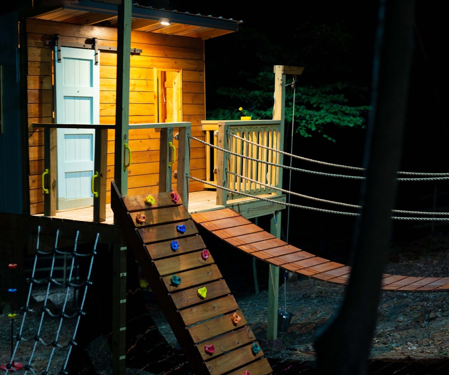 Outdoor tree house with mint green door and suspension bridge is illuminated with a downlight mounted above the porch.