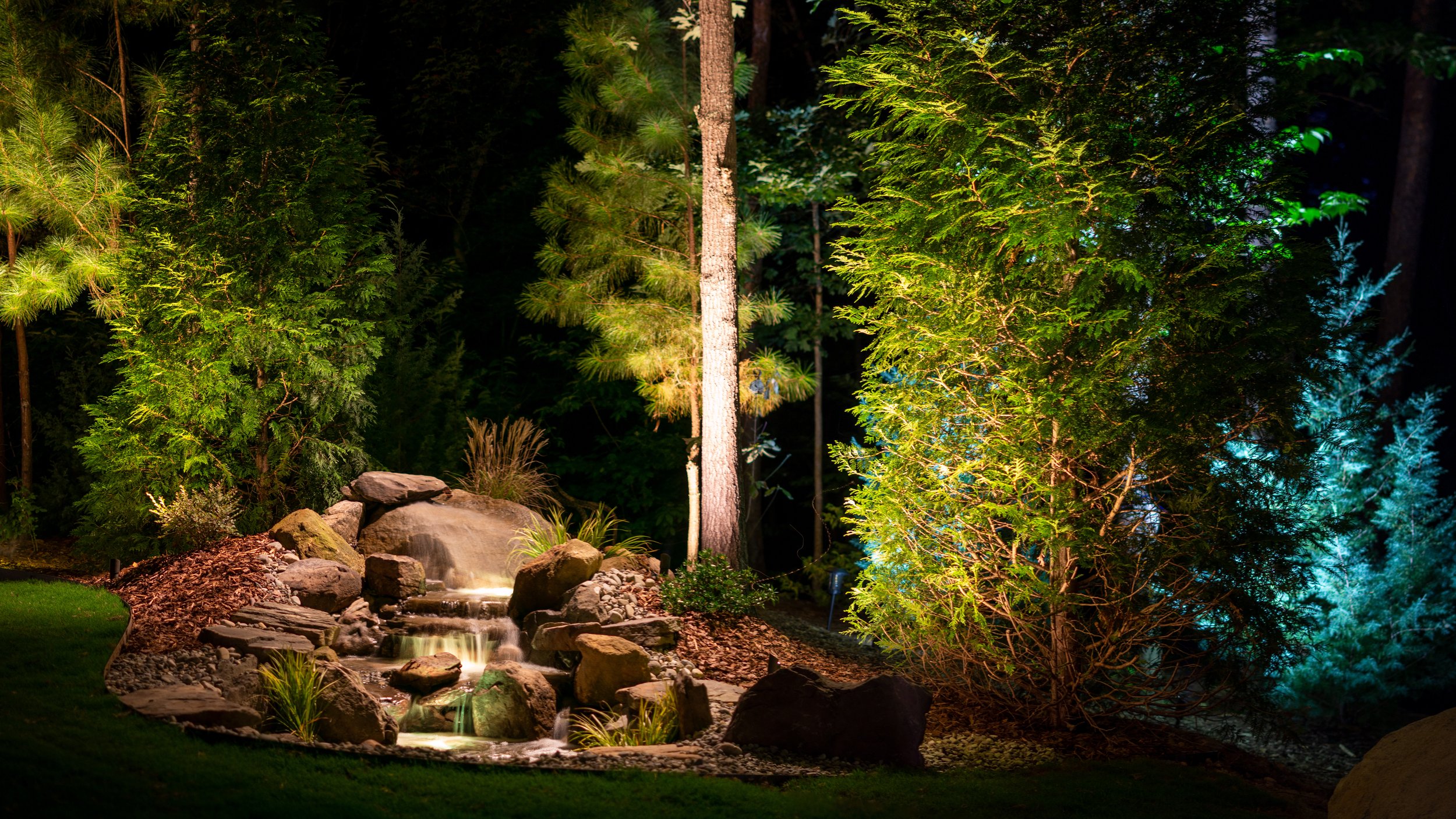 Water feature with a small waterfall surrounded by landscaping rocks is illuminated with underwater lights and down lighting. Surrounding trees are lit with tree accent lighting.