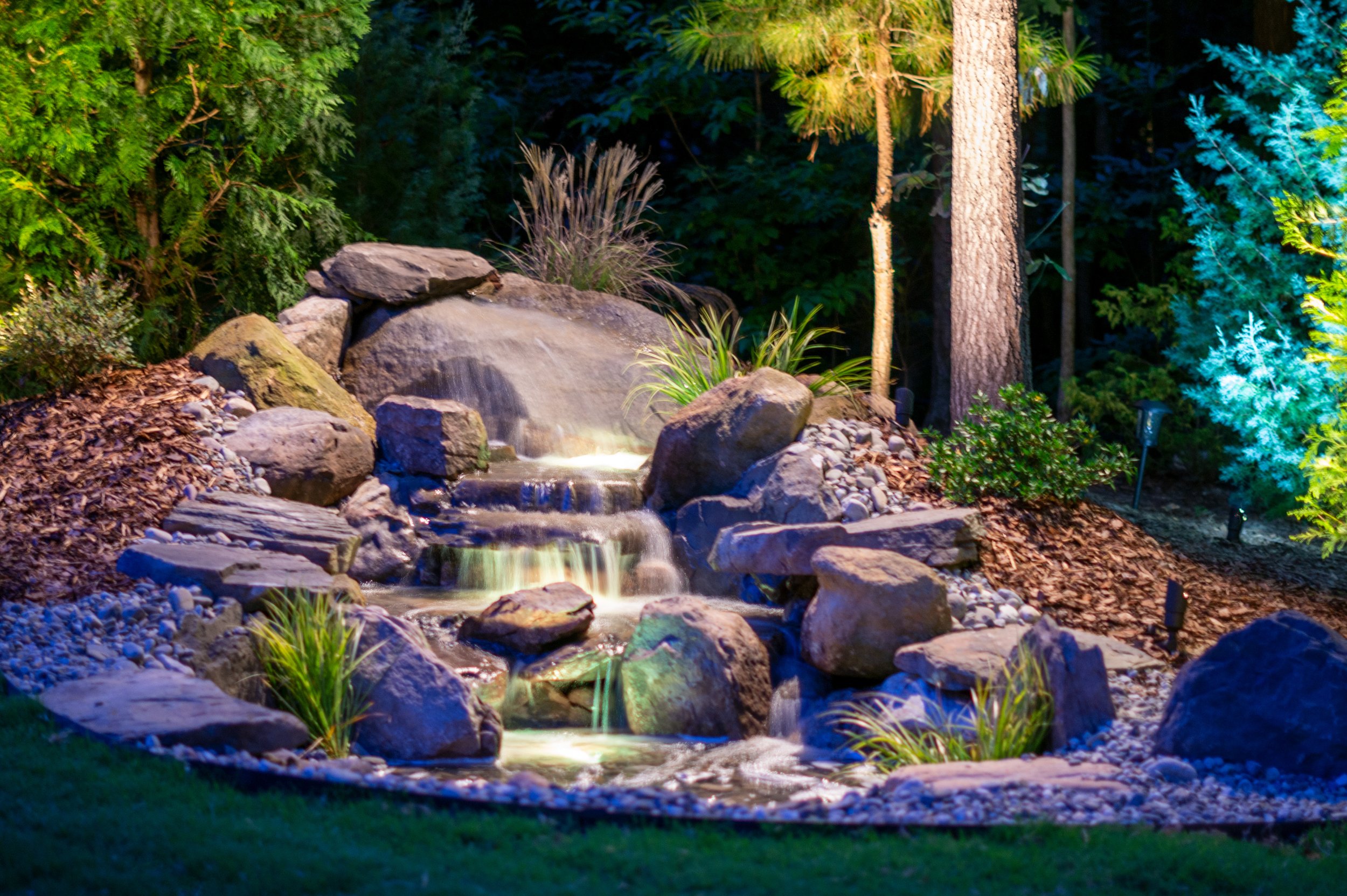 Close up image of a water feature with a small waterfall surrounded by landscaping rocks is illuminated with underwater lights and down lighting. Surrounding trees are lit with tree accent lighting.